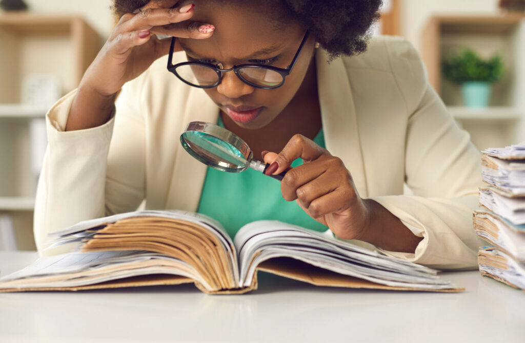 Closeup portrait of concentrated young african american woman auditor with magnifying glass scrutinizing financial documents at desk with paper folder pile in office. Investigation and audit checkup